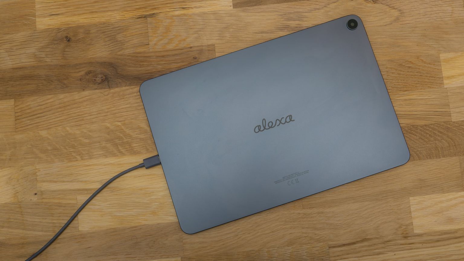 Rückseite des Alexa Tablet 12 Pro mit Alexa-Schriftzug und angeschlossenem USB-C-Kabel auf einem Holztisch
